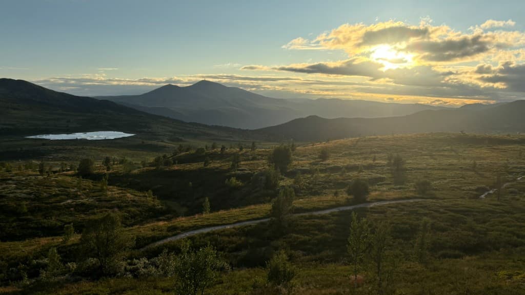 Sun breaking through clouds over highland hills, a lake, and a winding path