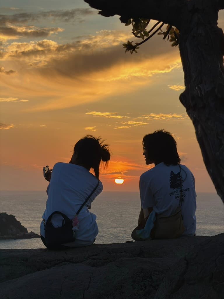 Two people sitting on a cliff watching the sun set over the sea